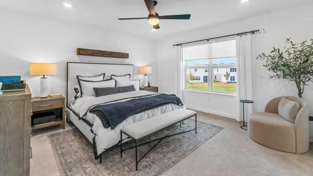 Bright, modern bedroom featuring a large bed with white and navy bedding, two matching bedside tables with lamps, and a soft area rug. A cushioned bench sits at the foot of the bed. To the right, a large window with white curtains allows natural light to fill the space. A round beige armchair, small side table, and decorative plant enhance the cozy and stylish atmosphere.
