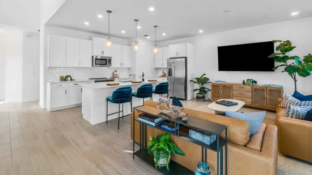 Bright, open-concept kitchen and living room featuring white cabinetry, stainless steel appliances, and a central island with three blue velvet barstools and pendant lighting above. The living area includes tan leather sofas, a modern coffee table, a large flat-screen TV mounted on the wall, and a wooden media console. A console table with books and a potted plant adds detail behind the sofa, and large potted plants bring natural elements into the space.
