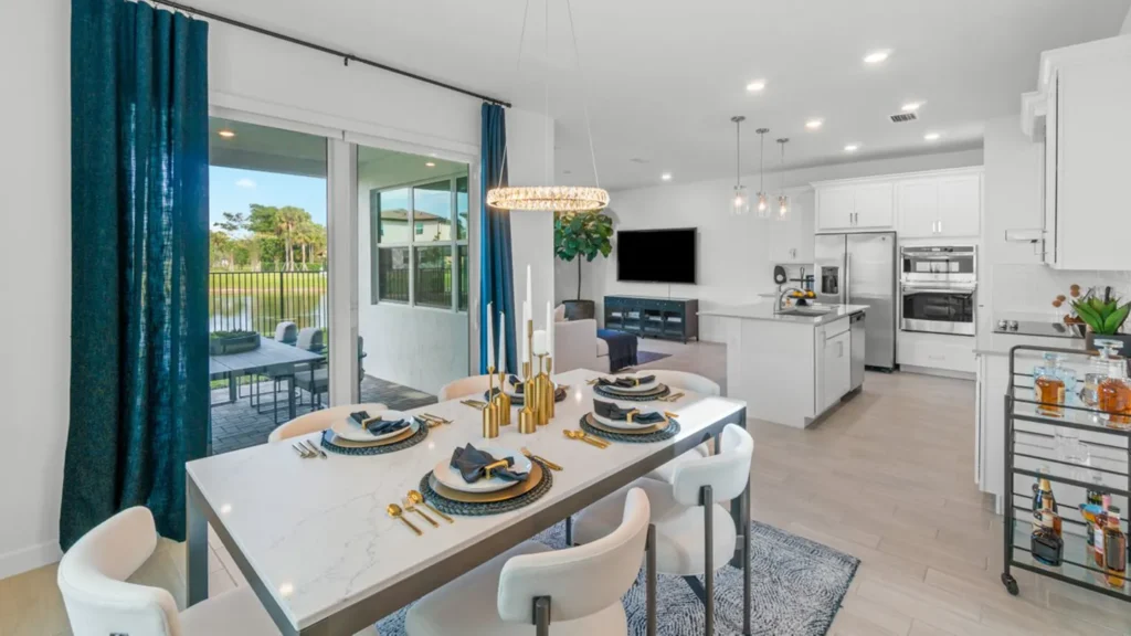 Elegant dining room with a marble-style rectangular table set for six, featuring gold flatware, black napkins, and tall modern candlesticks. To the left, large sliding glass doors open to a covered patio with outdoor seating and views of a pond and trees. In the background, a modern kitchen with white cabinets, stainless steel appliances, and a center island is visible, with pendant lights above and a bar cart stocked with drinks on the right.
