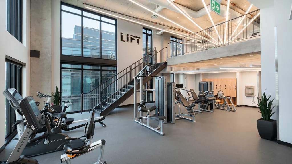 A bright, modern fitness center with floor-to-ceiling windows, housing various Matrix brand cardio and weight machines. Sleek linear lighting runs across the ceiling. Wooden lockers and a water station are visible in the background.