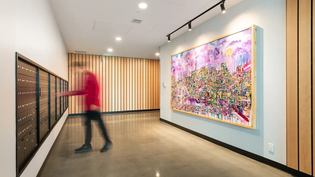 Modern building lobby featuring a row of black mailboxes on the left and a vibrant, colorful mural of a cityscape on the right wall. A person in a red sweater is walking past.