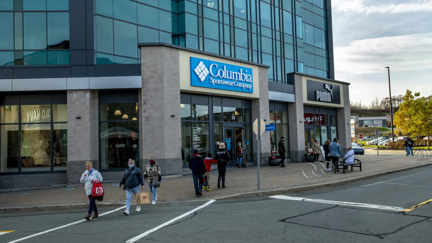 Street view of the Columbia Sportswear Company and PUMA outlet stores at Dartmouth Crossing, Nova Scotia. The modern building features glass panels and stone siding. A blue and white Columbia sign is above the main entrance, and a black and white PUMA sign is seen on the adjacent storefront. Several people walk in and out of the stores, some carrying shopping bags. A few shoppers are seated on benches near bike racks on the wide sidewalk, with a tree and cars in the background under a lightly overcast sky.