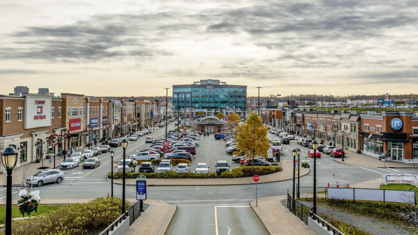Street-level view of Dartmouth Crossing’s retail area in Nova Scotia on a sunny day. The image shows a wide pedestrian-friendly street lined with parked cars and popular retail stores including Tommy Hilfiger, Skechers, and Lululemon on the left, and Life SalonSpa on the right. Shoppers are walking along the sidewalks, and a traffic worker in a safety vest is guiding cars. In the background, a glass office building rises at the end of the retail strip, with blue skies and scattered clouds overhead. The scene highlights a modern, open-air shopping district with landscaped walkways, benches, and street lamps.
