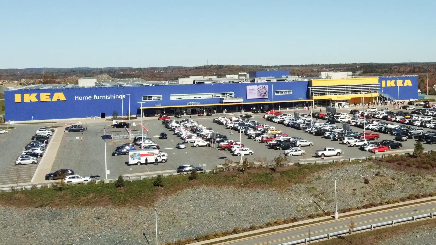 A wide-angle view of the large IKEA store at Dartmouth Crossing on a clear, sunny day. The building features the brand’s iconic blue and yellow color scheme, with large yellow “IKEA” signs and the words “Home furnishings” visible on the left side. The storefront is busy with a packed parking lot filled with cars and trucks. A U-Haul truck is parked near the entrance. The store has a modern, rectangular design with a digital billboard above the entrance and a secondary entrance on the right side. In the background, tree-covered hills and industrial buildings are visible under a pale blue sky.