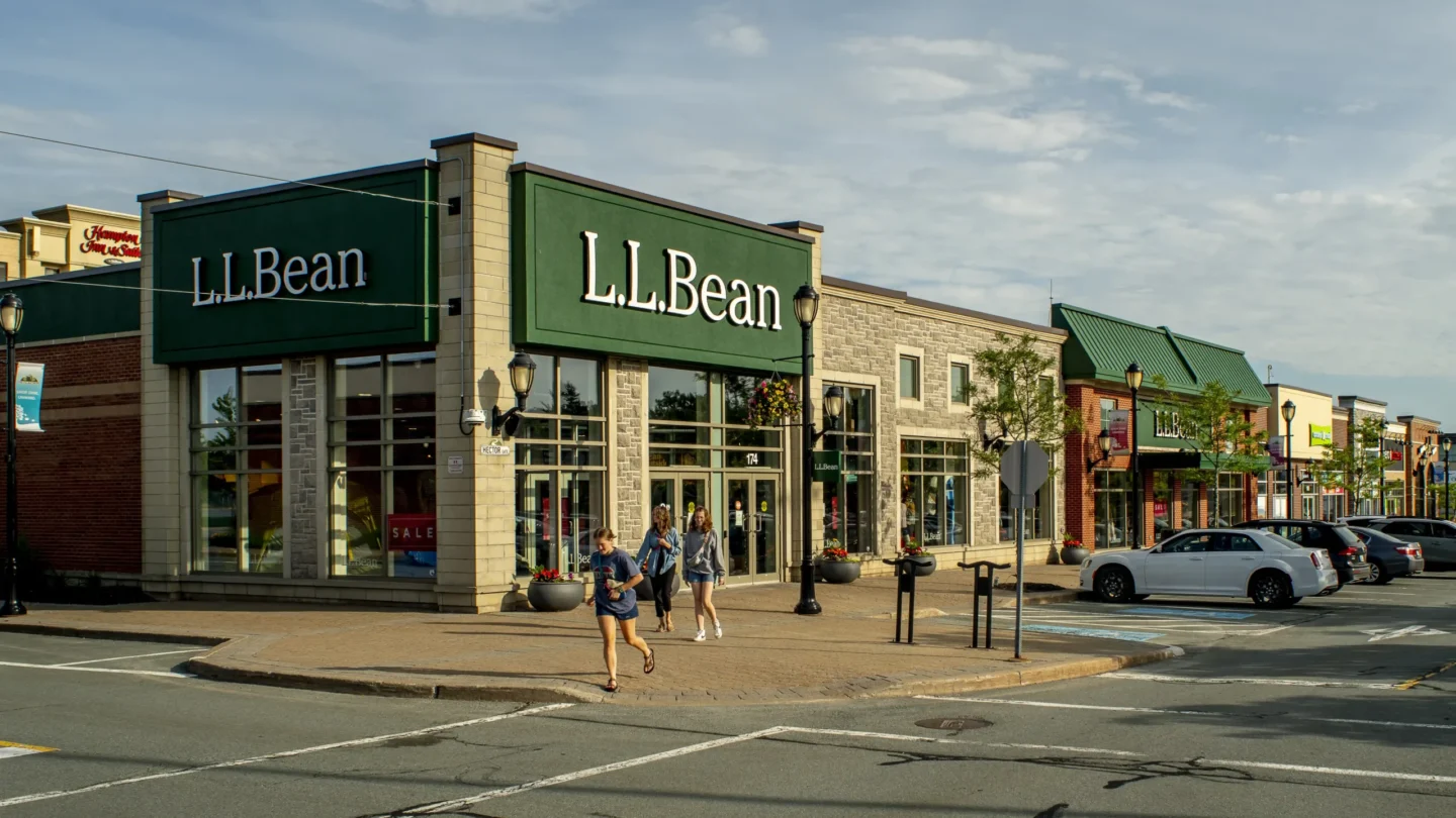 Daytime street-level view of the L.L.Bean retail store at Dartmouth Crossing in Nova Scotia. The store features large green signage and a stone-and-brick exterior with wide glass windows. Several people walk along the sidewalk outside the entrance, and planters with flowers and streetlamps decorate the pedestrian area. A white car is parked in an accessible parking space in front, and more storefronts with colorful signage line the street further down. The sky is partly cloudy, casting warm sunlight on the scene, suggesting a pleasant shopping environment.