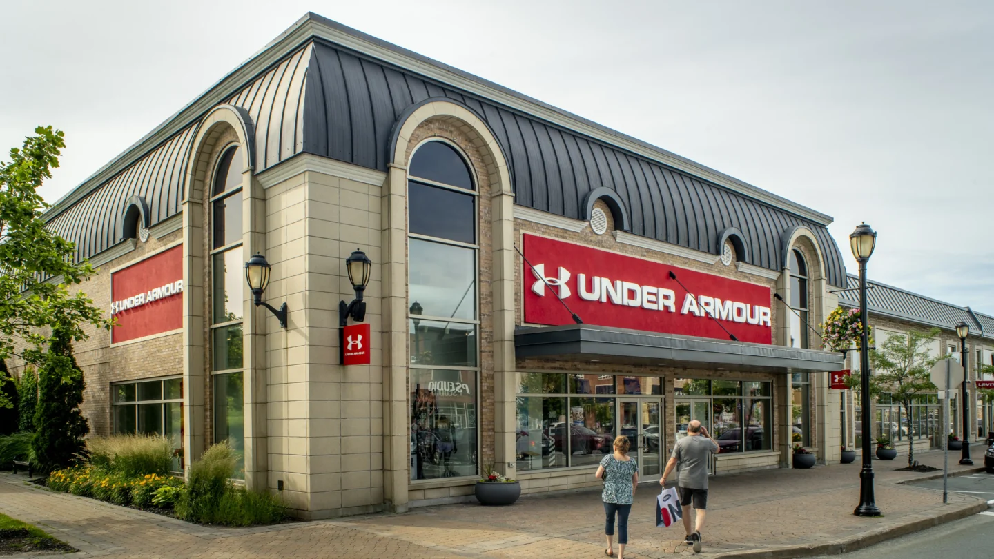 Street-level view of the Under Armour store at Dartmouth Crossing in Nova Scotia. The building has a tan and beige exterior with large arched windows and a black metal roof. Prominent red signs with the Under Armour logo are mounted on the front and side of the building. A pair of people walk on the wide sidewalk toward the entrance, one carrying a shopping bag. Flowering plants, streetlamps, and hanging baskets line the walkway, creating an accessible and welcoming shopping environment under a lightly overcast sky.