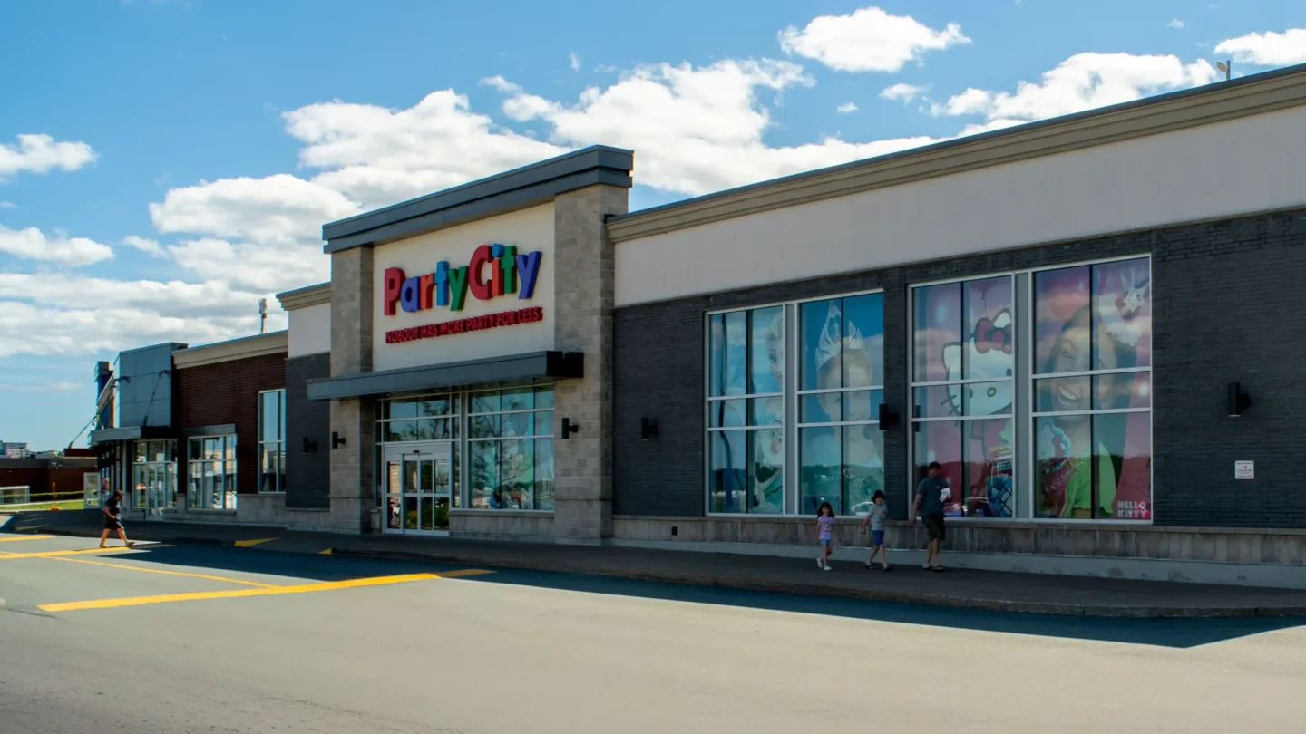 Exterior view of the Party City store at Dartmouth Crossing on a bright day with a partly cloudy sky. The store’s sign is prominently displayed above the entrance, with colorful letters spelling “Party City” and the slogan “Nobody Has More Party for Less” in red below. Large windows on the right display themed images, including Hello Kitty and festive decorations. A man walks with two children on the sidewalk in front of the store, while another person crosses the drive lane on the left. The building exterior is a mix of beige stone, white walls, and dark gray brick.