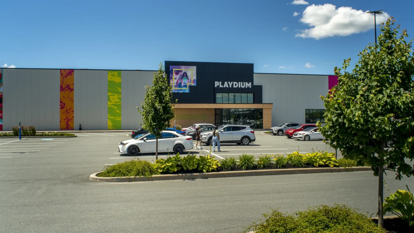 Exterior view of the Playdium entertainment complex in Dartmouth Crossing on a bright, sunny day. The modern building has colorful vertical accent panels and a dark blue sign with "PLAYDIUM" written in bold white letters above the main entrance. A large digital screen above the entrance displays an image of people enjoying an activity. Several cars are parked in front, with two people walking through the lot. Well-maintained trees and shrubs line the parking area under a clear blue sky with a few white clouds.