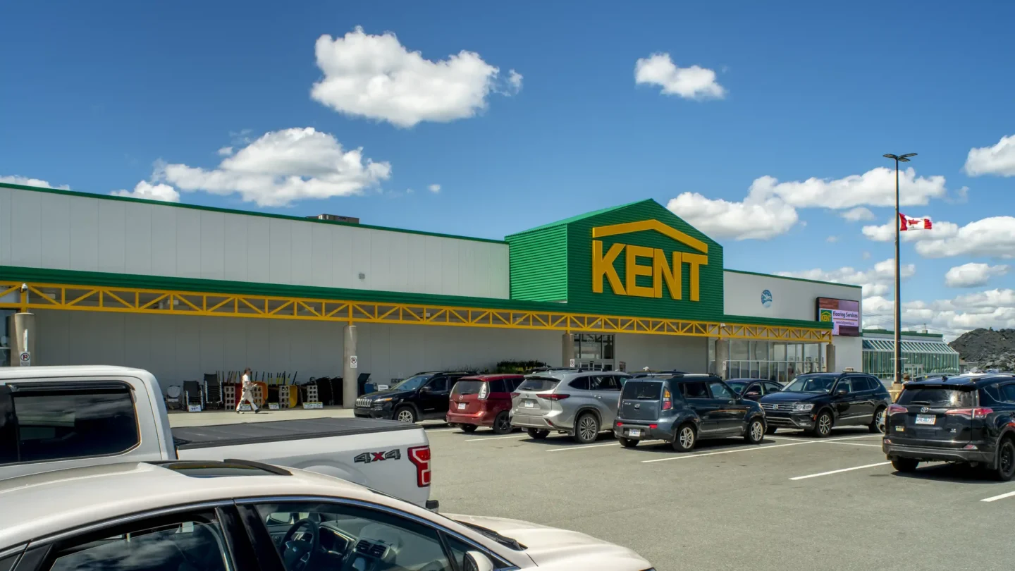 Exterior view of the Kent Building Supplies store at Dartmouth Crossing on a sunny day with a bright blue sky and scattered white clouds. The large building features the Kent logo in bold yellow letters on a green facade, with a yellow and green awning stretching across the front. The parking lot in the foreground is partially filled with various vehicles, including cars, SUVs, and trucks. A Canadian flag is visible on a tall pole to the right, and some outdoor merchandise such as chairs is displayed along the side of the store.