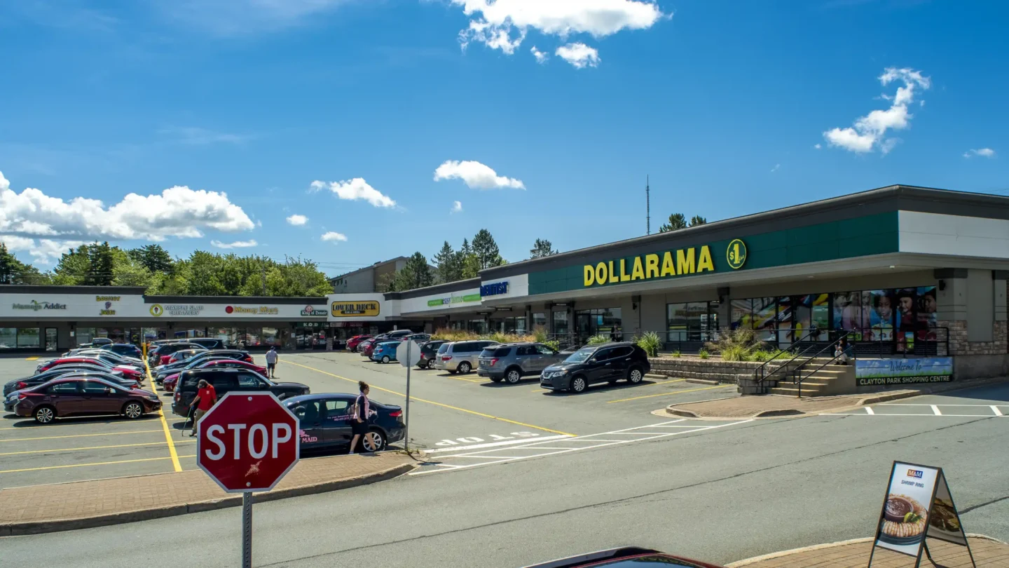 A sunny, wide-angle view of the Clayton Park shopping plaza focused on the Dollarama store. To the left of Dollarama are other storefronts including a dentist office, easyhome, The Lower Deck, K.O. Donair, and Massage Addict. Several parked cars and a few pedestrians are visible in the foreground, with a clear blue sky and scattered clouds overhead. A “STOP” sign and a promotional sandwich board stand near the curb in front of the parking area. Trees and low-rise buildings form the backdrop.