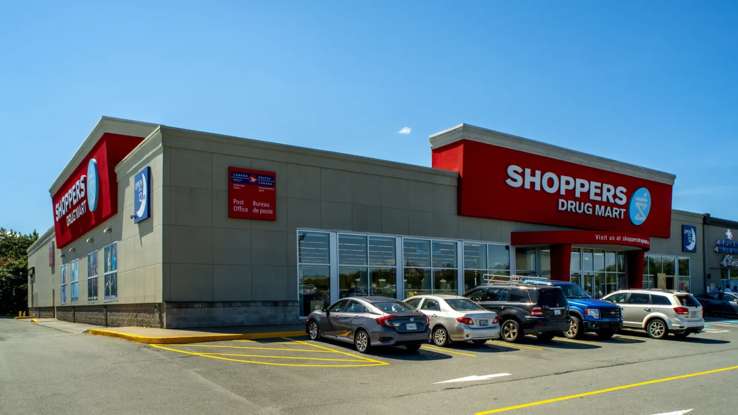 A bright, sunny view of the Shoppers Drug Mart at Clayton Park Shopping Centre, featuring a bold red storefront with large signage and a Canada Post outlet sign mounted on the side. Several cars are parked in front of the building, and the entrance is marked by red columns and a glass façade. A sign indicates the store is open 24 hours. Clear blue sky overhead with no visible clouds.