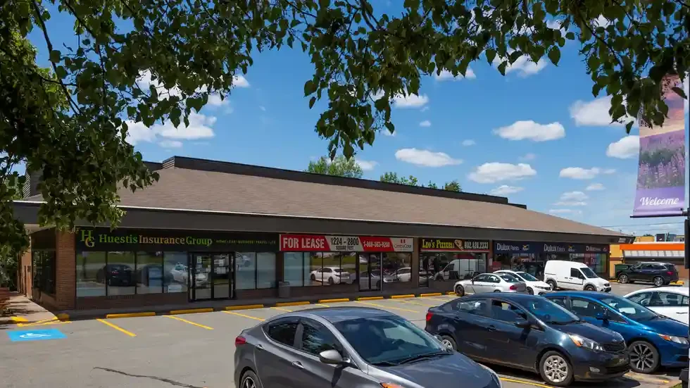 Street-level view of Bedford Highway Centre, a single-storey commercial building with signage for tenants such as Huestis Insurance Group and Dollarama, visible storefronts, accessible parking, and several parked vehicles in front.
