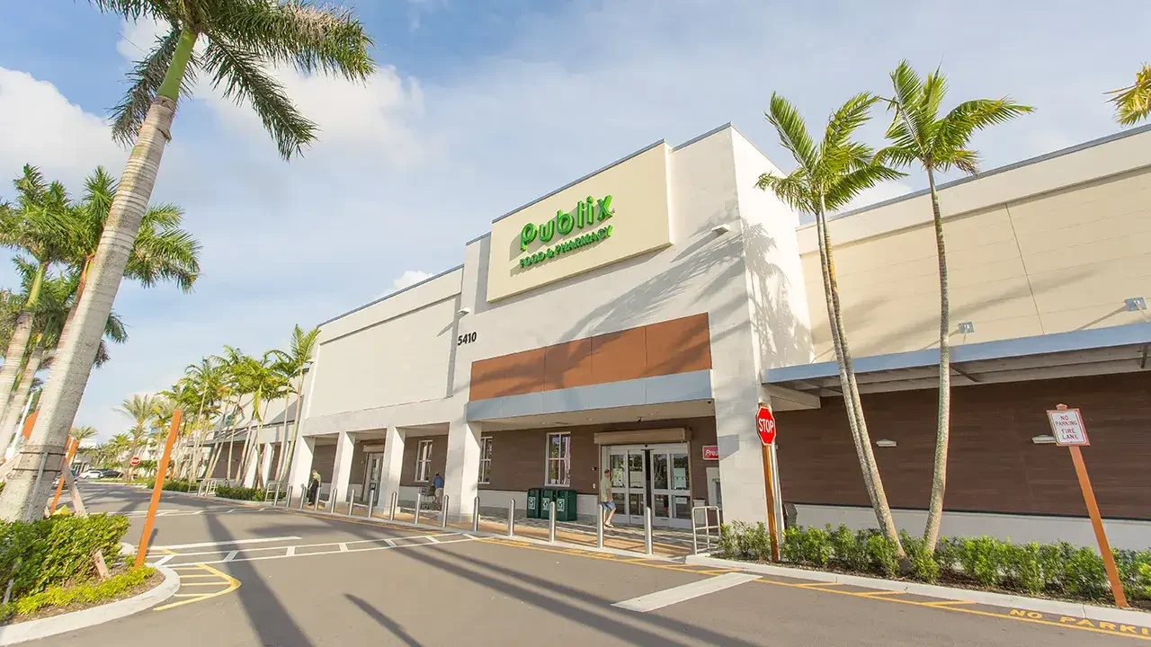 Exterior view of a Publix Food & Pharmacy storefront at 5410 Alton Road in Palm Beach Gardens, Florida. The modern building features a clean, beige and white façade with a prominent green Publix sign above the entrance. The surrounding area includes palm trees, a pedestrian walkway, and a clearly marked stop sign near the front of the store. The weather is sunny with a partly cloudy sky.