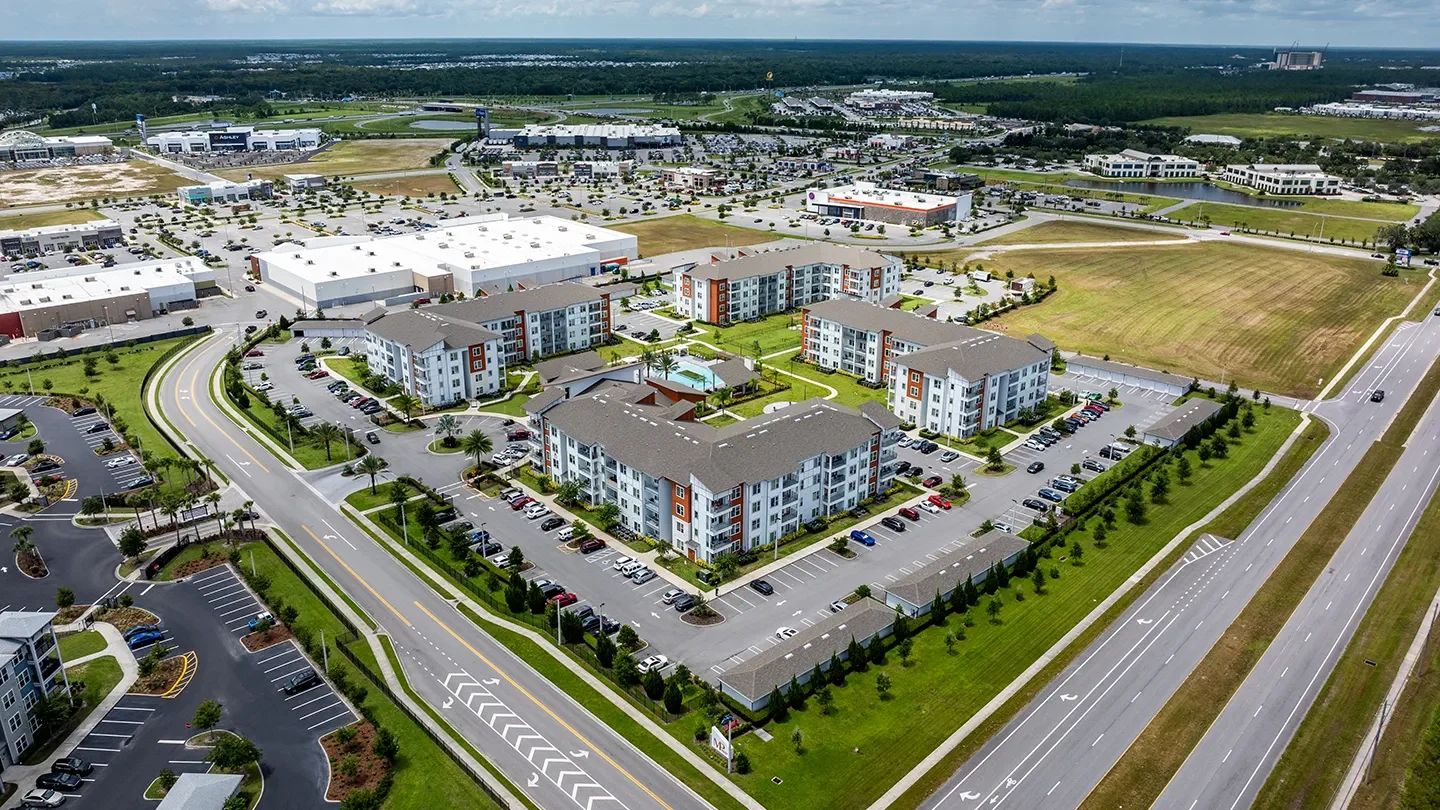 Aerial view of Madison Pointe Apartments in Daytona Beach, Florida. The image shows a cluster of modern four-story apartment buildings with white facades and red-orange accents. The buildings are arranged around a central courtyard that includes a swimming pool and landscaped green space. The property is surrounded by parking lots and bordered by wide multi-lane roads. In the background, large retail stores, commercial buildings, and a network of access roads and green spaces are visible, with wooded areas and distant structures lining the horizon.