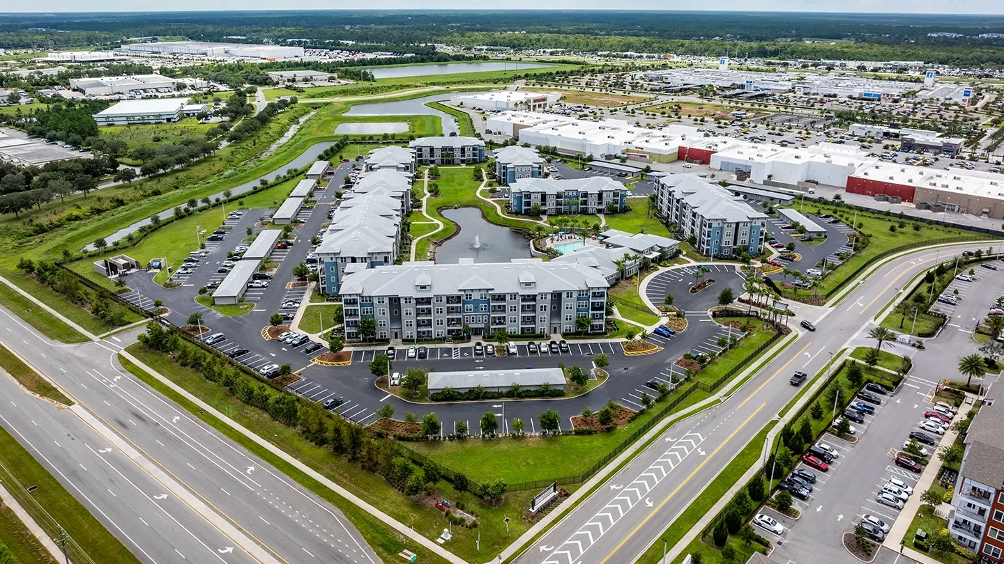 Aerial view of Tomoka Pointe Apartments in Daytona Beach, Florida. The image features multiple modern four-story apartment buildings arranged around a central landscaped courtyard with a large pond and fountain. The buildings are painted in neutral tones with dark grey roofing. Curved walking paths and driveways wind through the complex, which also includes covered parking areas and neatly landscaped green spaces. In the background, large white-roofed retail buildings and parking lots of the Tomoka Town Center are visible, surrounded by roads, waterways, and patches of greenery.