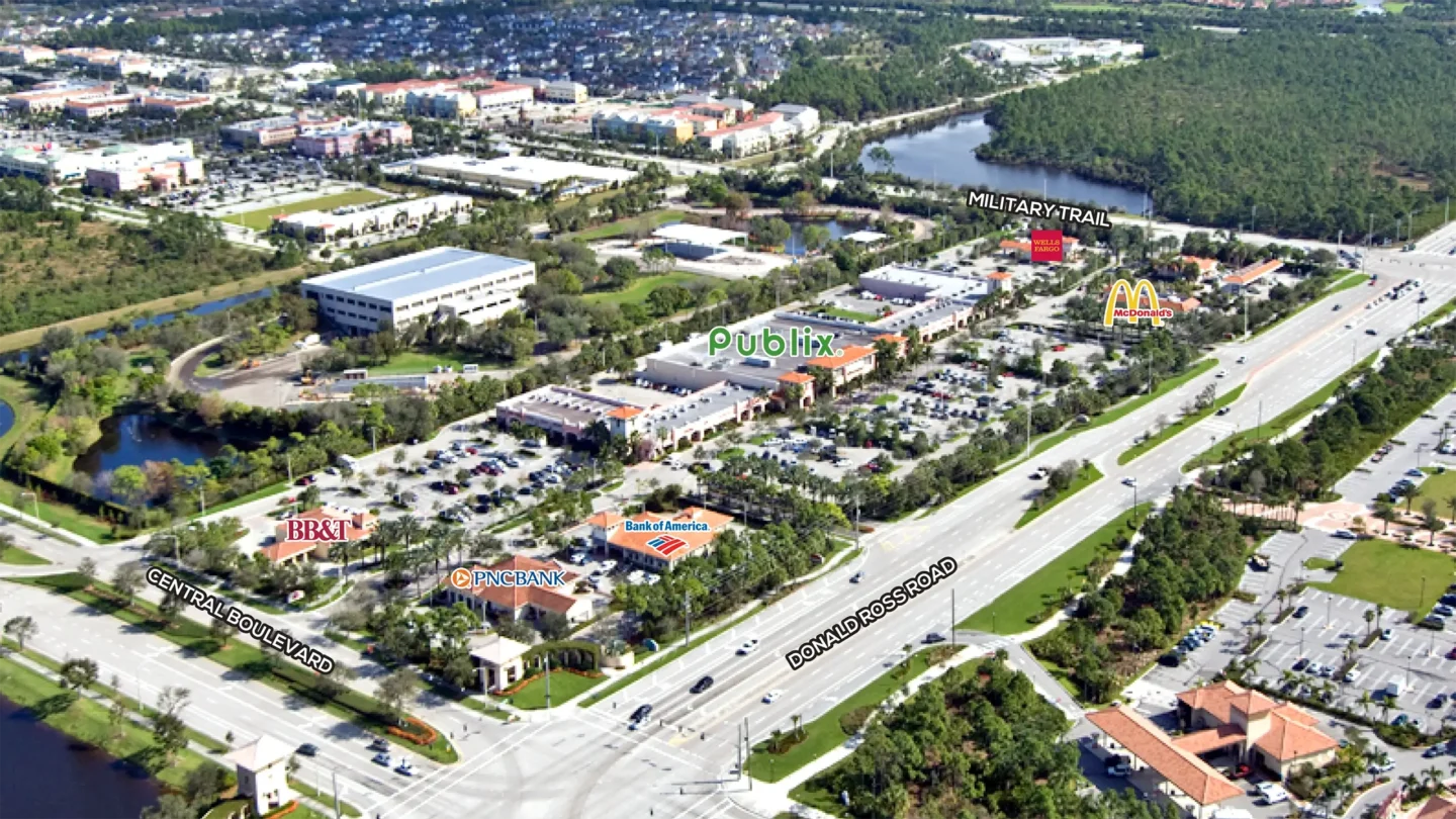 Aerial photograph of Abacoa Plaza at the intersection of Donald Ross Road and Central Boulevard in Jupiter, Florida. Major tenants are labeled, including Publix, McDonald’s, Wells Fargo, BB&T, PNC Bank, and Bank of America. The plaza includes several buildings surrounded by palm trees, landscaped parking lots, and nearby roads and waterways.