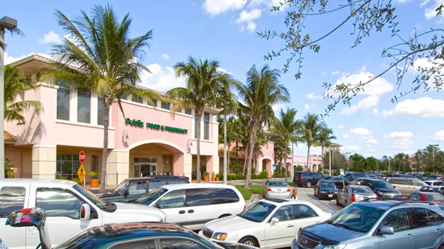 Street-level image of the Publix supermarket at Abacoa Plaza with its main entrance centered under an arch. The parking lot in the foreground is filled with vehicles, and palm trees and tropical landscaping surround the building. The sky is mostly clear with scattered clouds.