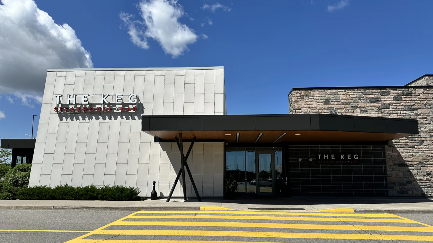 A straight-on view of The Keg Steakhouse + Bar entrance at Park Place shopping centre. The modern exterior features a tall light grey tiled facade with bold signage reading “The Keg Steakhouse + Bar” in red and black letters. A wide black awning with wooden paneling underneath extends over the double-door entrance. The right side of the building is clad in textured stone. A bright yellow accessible crosswalk is painted on the asphalt in front, leading directly to the entrance. The sky is clear and sunny with a few white clouds.