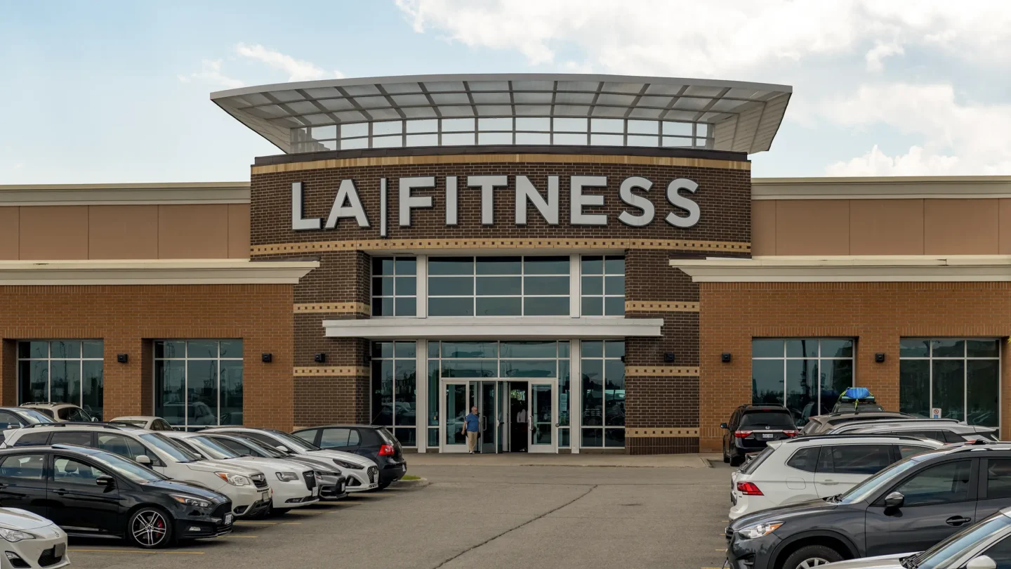 A frontal view of the LA Fitness gym entrance at Park Place shopping centre. The building features a large, curved brick facade with bold white letters spelling "LA FITNESS" above a glass entryway. A modern, curved metal canopy tops the structure. The lower level of the building has large glass windows and double glass doors. The foreground shows a full parking lot with various parked cars, and one person exiting the facility. The sky is partly cloudy with soft light.