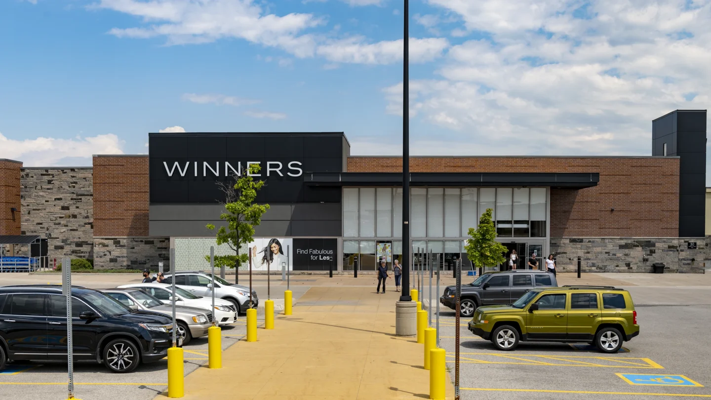 A clear daytime view of the exterior of the Winners retail store at Park Place shopping centre. The building features a modern facade with black and brick accents, along with a stone base. Large vertical windows frame the entrance, and a branded sign above reads “WINNERS.” In the foreground, a row of parked vehicles is visible, including accessible parking spots marked with bright yellow paint. Several people are seen walking near the entrance and through the parking area. The sky is partly cloudy with blue patches visible.