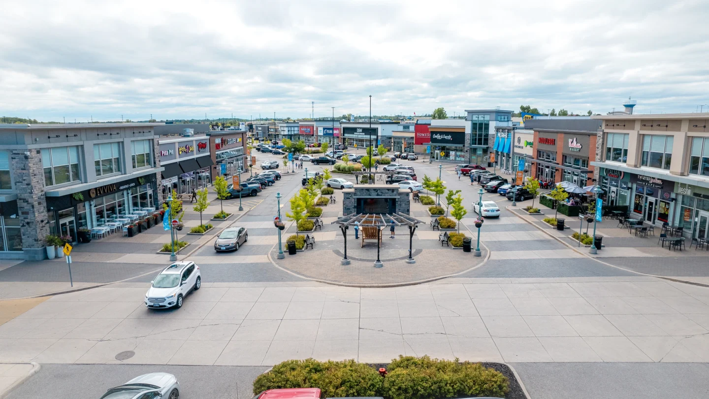A daytime street-level view of Park Place Shopping Centre showing a central pedestrian plaza with a covered seating area and symmetrical rows of young trees and benches. On both sides of the plaza, there are modern retail buildings with ground-level shops and restaurants. Visible storefronts include Evviva Breakfast & Lunch, Pita Pit, Booster Juice, The Chopped Leaf, The Beer Store, Telus, DSW, HomeSense, Carter’s OshKosh, Skechers, Smoke’s Poutinerie, Italian Eatery, and Freshii. Several parked cars line both sides of the main walkway, and a few people are walking or sitting outdoors. The sky is overcast, contributing to a cool, evenly lit atmosphere.