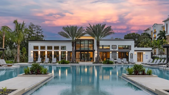 Luxury NUVO Club pool area at sunset with palm trees, lounge chairs, and a modern clubhouse reflecting in the water. The sky displays soft hues of pink and purple.