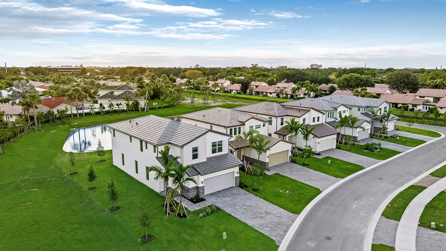 Aerial view of NUVO Boca featuring rows of two-story white homes with gray roofs, manicured lawns, palm trees, and a small pond surrounded by green space. A curving paved road runs through the quiet residential area under a partly cloudy sky.