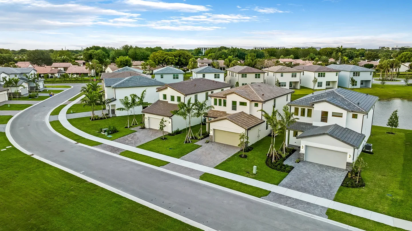 Aerial view of NUVO Boca street lined with modern two-story white homes featuring tile roofs, green lawns, and palm trees. The neighborhood is bordered by a pond and surrounded by additional residential areas with lush greenery and a blue sky overhead.