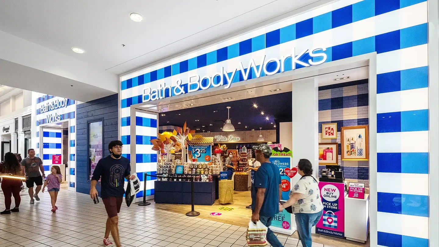 Entrance to Bath & Body Works, with a blue and white checkered design framing the store. Inside, displays of seasonal products and signs promoting sales are visible, including a table with $3.25 items and promotional signs for buy-online, pick-up in-store. Shoppers are walking past the entrance, some carrying shopping bags.