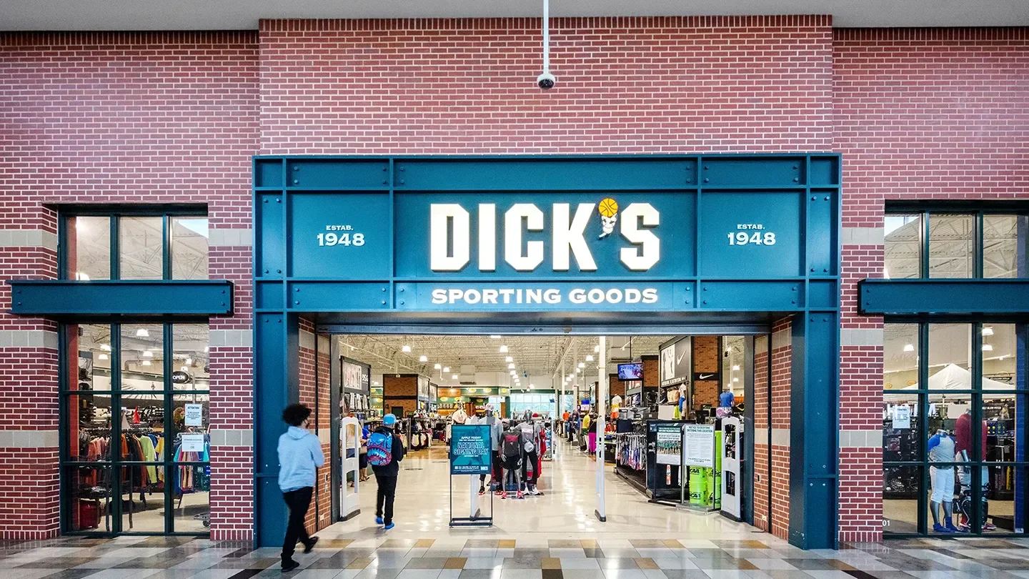 Front view of Dick’s Sporting Goods, featuring a bold green sign with large white lettering and a basketball graphic as part of the logo. The store has a red brick facade and large windows on either side, revealing displays of athletic clothing and equipment. Several shoppers are entering the well-lit interior.