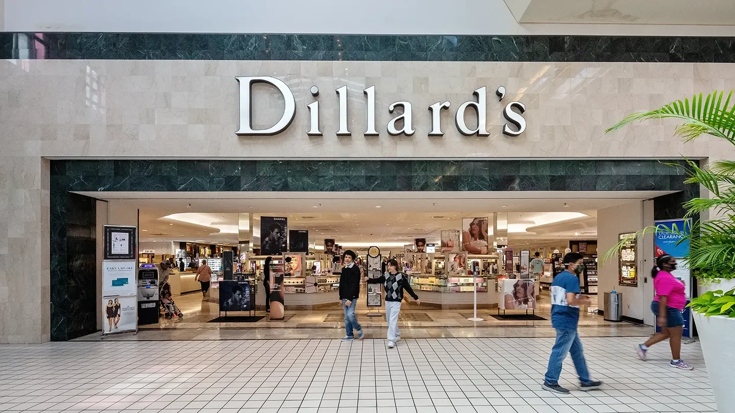 Entrance of Dillard’s department store with large signage against a polished stone wall. The interior cosmetics section is visible with product displays and promotional posters. Shoppers walk past and inside the store.