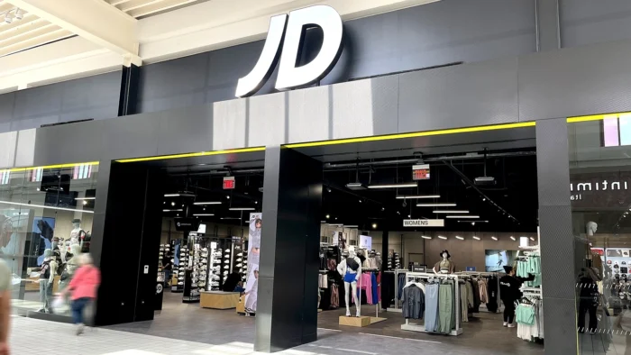 Modern storefront of JD Sports with large white “JD” signage above black-framed entrances. Inside, mannequins and clothing racks are neatly arranged under bright lights. A few shoppers browse near the entrance.