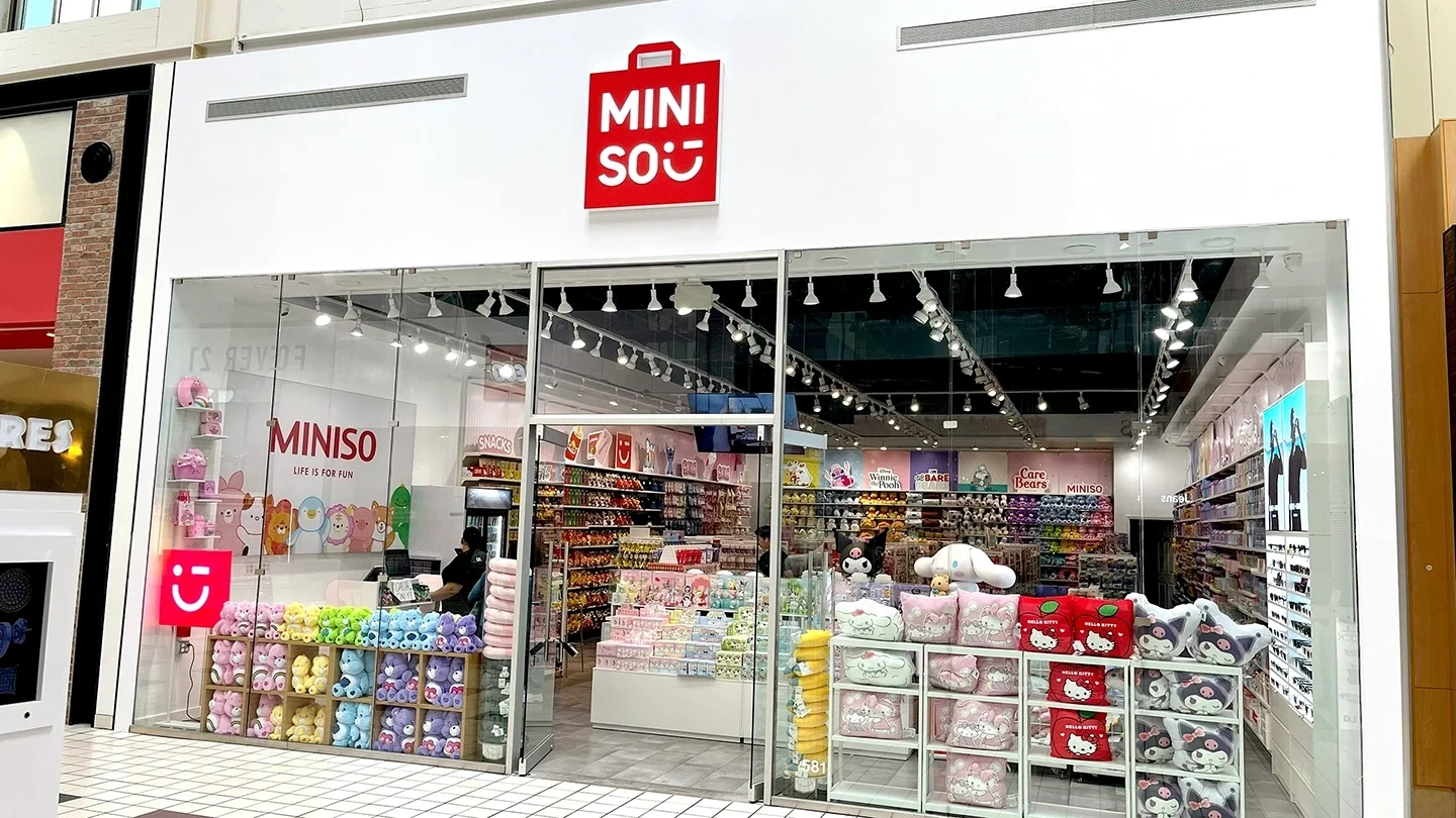 Exterior view of the MINISO store in a mall, with large glass windows displaying colorful plush toys, cosmetics, and character-themed merchandise like Hello Kitty and Care Bears. A cashier is visible inside the brightly lit store under a red sign that reads 