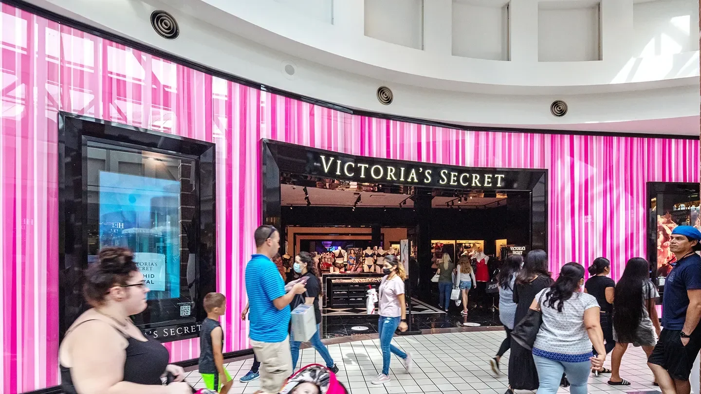 A crowd of shoppers walks past the brightly lit Victoria’s Secret storefront in a mall. The store features a distinctive pink striped wall and black-framed entrance. People of all ages are seen walking and browsing.
