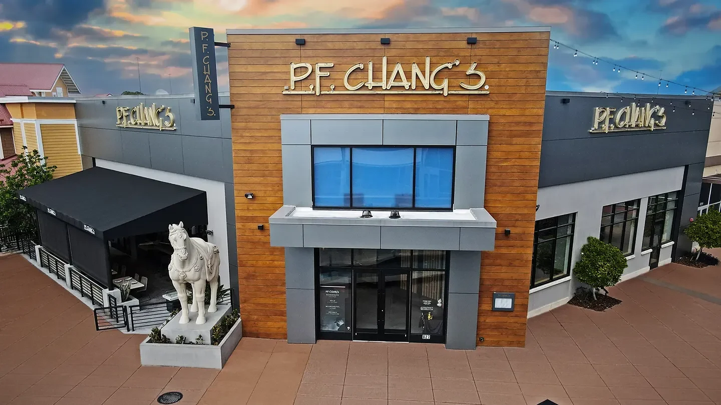 Front exterior of P.F. Chang’s restaurant during dusk, featuring modern architecture with wood paneling and grey accents. A large white horse statue is positioned at the entrance next to an outdoor dining area covered by a black awning."
