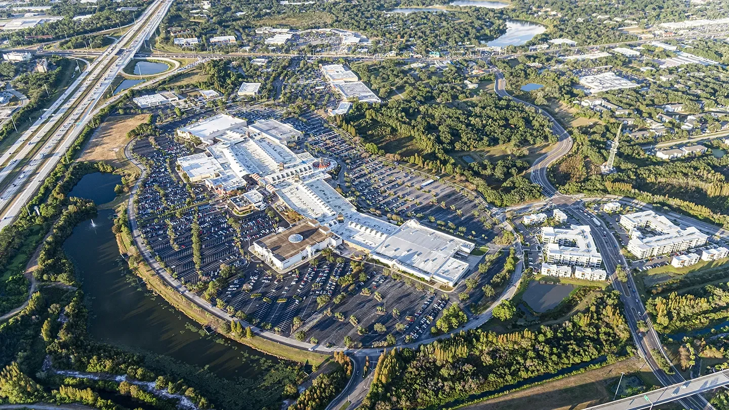 Aerial view of the Brandon Exchange shopping center, showcasing a large, curved commercial complex with expansive white-roofed buildings and surrounding parking lots. The site is densely populated with cars and encircled by natural features, including small lakes, wooded areas, and nearby roadways, with a major highway interchange visible in the upper left. Adjacent residential and commercial developments appear in the background.