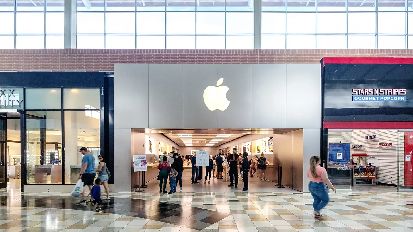 Shoppers walking past the Apple Store. The store features a minimalist gray facade with a glowing Apple logo above the entrance. Inside, people interact with products at wooden tables.
