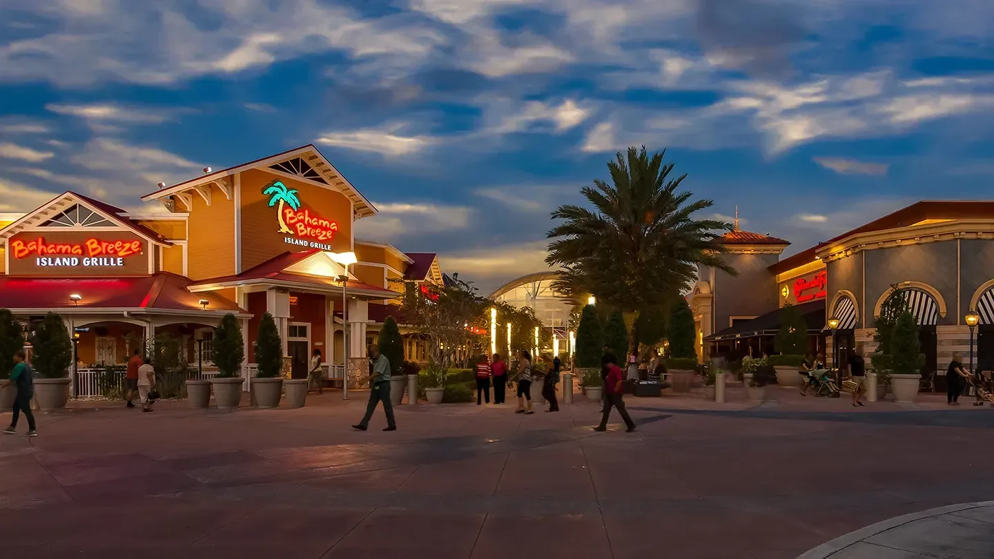 Evening view of Bahama Breeze Island Grille, a tropical-themed restaurant with warm yellow and red exterior tones, white trim, and a neon sign with a palm tree graphic. The restaurant is surrounded by large potted plants and palm trees. People walk casually along the wide, well-lit plaza under a dramatic blue and orange sunset sky. Additional buildings and storefronts, including another restaurant, are visible in the background.