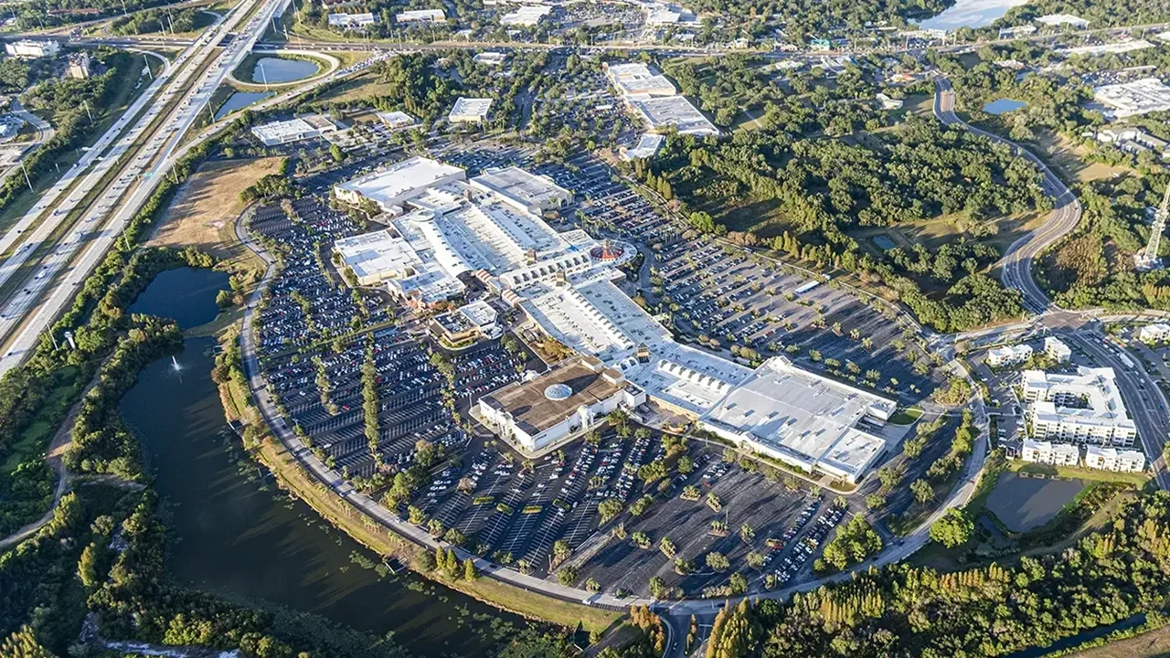 Aerial view of the Brandon Exchange shopping center, showcasing a large, curved commercial complex with expansive white-roofed buildings and surrounding parking lots. The site is densely populated with cars and encircled by natural features, including small lakes, wooded areas, and nearby roadways, with a major highway interchange visible in the upper left. Adjacent residential and commercial developments appear in the background.