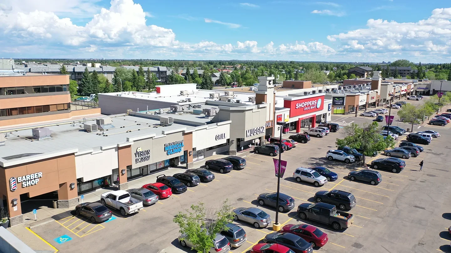 Ground-level view of the Marketplace at Callingwood with several storefronts including a barber shop, Vision by Design Optometry, a medical clinic, MUGGNZ and Shoppers Drug Mart. The foreground shows a parking lot with numerous vehicles and pedestrians under a sunny sky with scattered clouds.