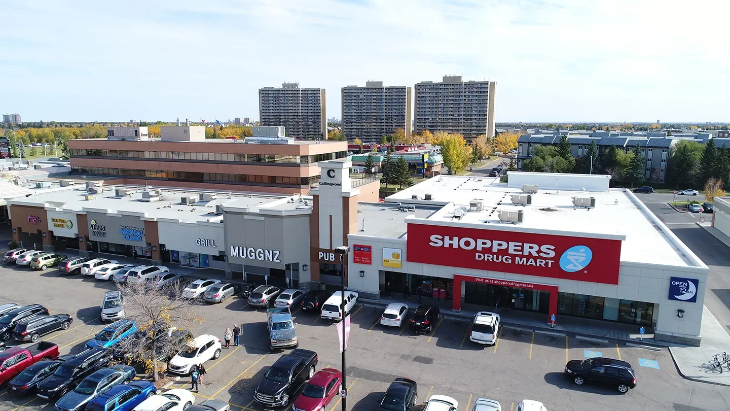A wide view of a shopping plaza at Marketplace at Callingwood featuring Shoppers Drug Mart in the foreground. Adjacent storefronts include MUGGNZ, Vision by Design Optometry, and other local businesses. The parking lot is full of cars, and tall apartment buildings can be seen in the background under a clear sky.