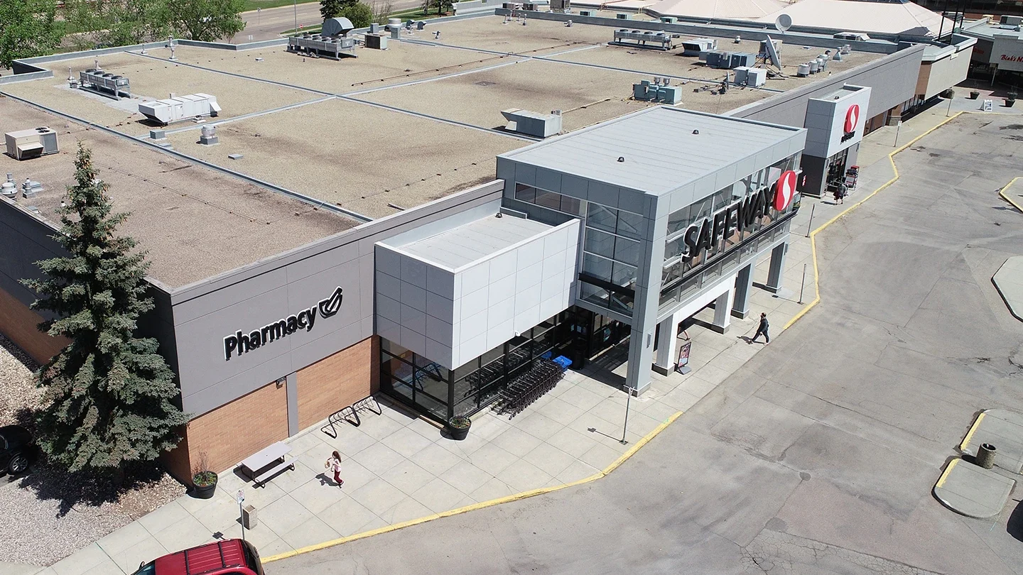 Aerial view of a Safeway grocery store with a modern grey and glass entrance and an adjacent pharmacy section. The storefront includes shopping cart storage and sidewalk access, with a few people walking near the entrance. The surrounding area includes a large parking lot and minimal landscaping with a tree and planter.