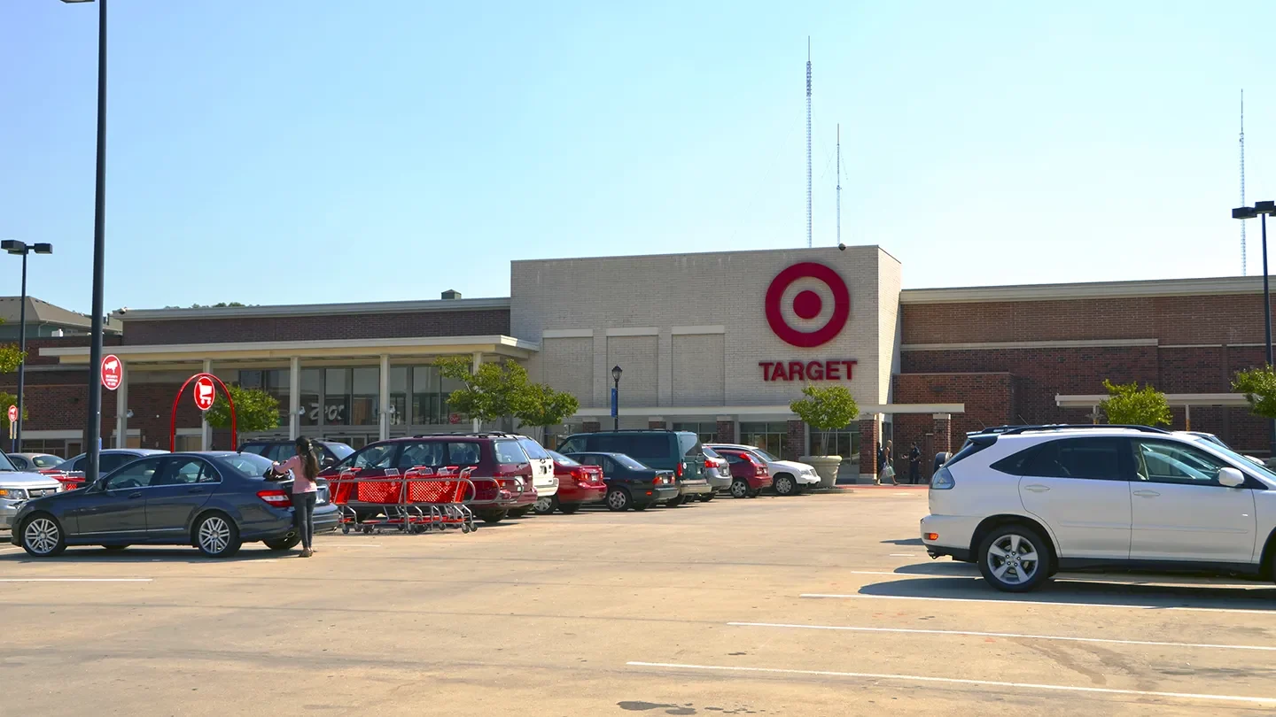 Daytime view of the Target store entrance at the Edgewood Retail District in Atlanta, Georgia. The building is made of red and white brick, with the large red Target logo and name displayed above the entrance. In front of the store, several cars are parked in the lot, and red shopping carts are stacked near a cart return station. A woman is seen placing items into the trunk of a dark-colored sedan. The sky is clear and blue, and small trees line the edge of the lot.