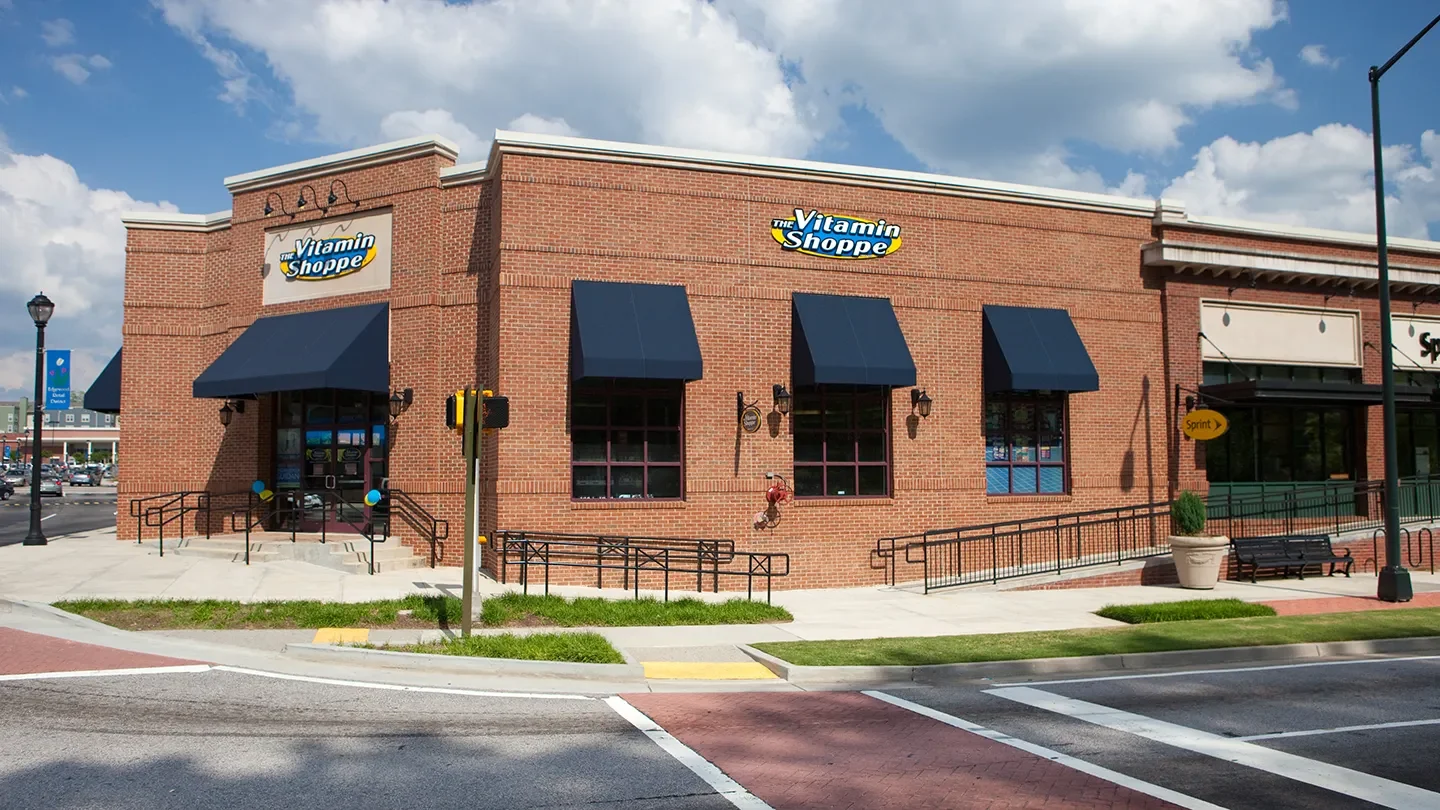 Exterior view of The Vitamin Shoppe in the Edgewood Retail District in Atlanta, Georgia. The store is housed in a red brick building with three navy blue awnings over the windows and a matching entrance canopy. A ramp and stair railings provide accessible entry, and a traffic signal is located at the corner in front of the building. The storefront also features a neighboring Sprint store on the right. The sky is partly cloudy with blue skies peeking through.