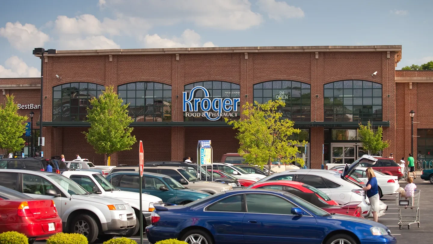 Exterior view of the Kroger Food & Pharmacy store located in the Edgewood Retail District in Atlanta, Georgia. The store features a large red brick facade with tall, arched windows and a blue and white Kroger sign above the entrance. The entrance doors are located beneath the windows, and a sign on the window reads “24 OPEN.” In the foreground, the parking lot is filled with cars, and people are seen walking or unloading groceries, including a woman and child near a shopping cart.
