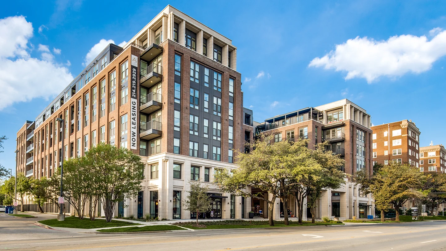 A multi-story modern apartment building with a brick and stone facade, large windows, and balconies. A vertical banner reads "Now Leasing" with a phone number. Mature trees and a sidewalk line the street in front of the building, with a bright blue sky overhead.