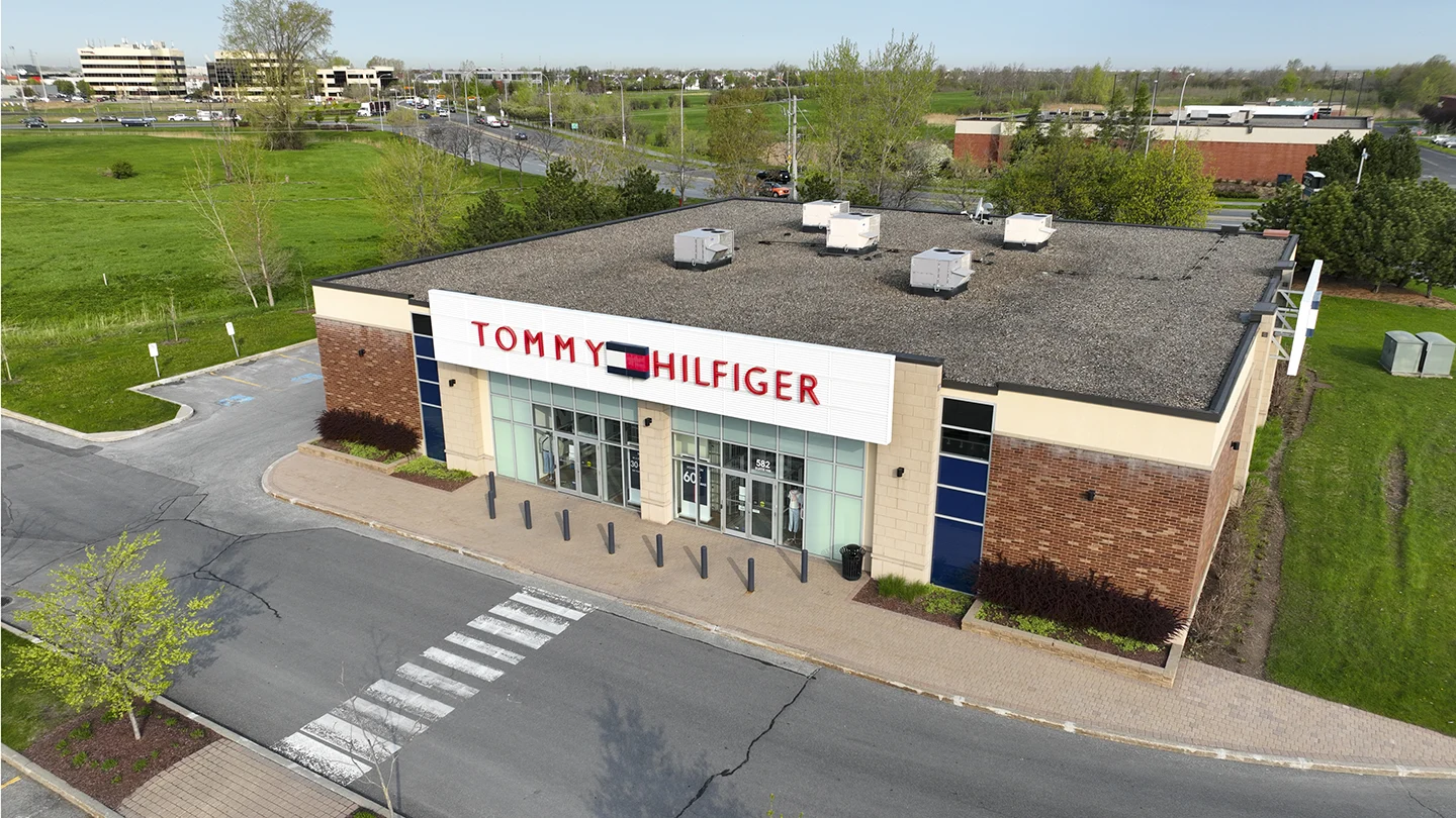 Aerial daytime view of a Tommy Hilfiger retail store located on a corner lot surrounded by green space and intersecting roads. The storefront features a large white sign with red and navy blue lettering spelling “TOMMY HILFIGER” above a row of large glass windows and a double glass entrance door labeled “582.” A brick pathway runs in front of the store, with several black bollards and a trash bin near the entrance. Visible on the windows are promotional posters, including a “60%” discount sign. The building exterior combines beige stone and red brick with accessible parking spaces to the left.