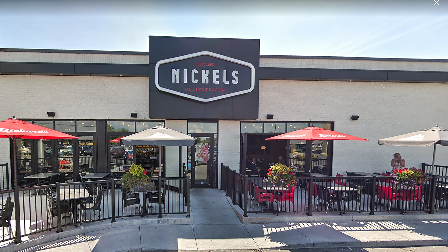 Street-level view of the Nickels Delicatessen restaurant exterior at Les Jardins Dorval. The facade features a prominent black and white Nickels sign with red text reading "Est. 1990" and "Delicatessen." The front patio includes black metal fencing, black dining tables, and umbrellas in red and grey with “Rickard’s” branding. Planters with colorful flowers decorate the patio. One woman is standing near the right edge of the patio. The restaurant interior is partially visible through large glass windows.