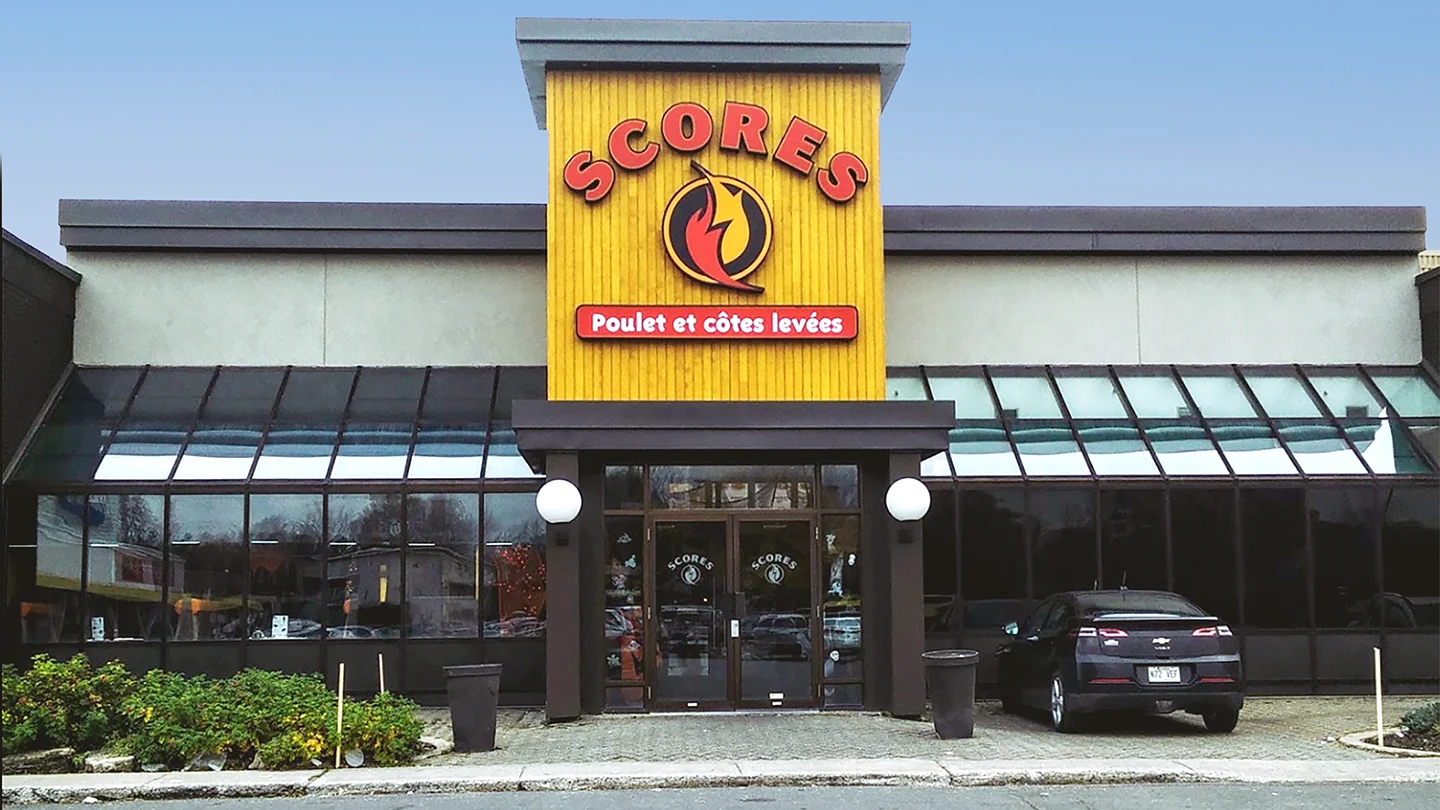 Front view of a Scores restaurant located at Les Jardins Dorval. The building features a prominent yellow vertical panel in the center with the red Scores logo and a stylized flame icon. Below the logo is a red sign with white text that reads “Poulet et côtes levées,” indicating the restaurant specializes in chicken and ribs. The entrance includes double glass doors with round white wall lamps on either side. A black car is parked directly in front of the restaurant entrance, and large glass windows line the facade.