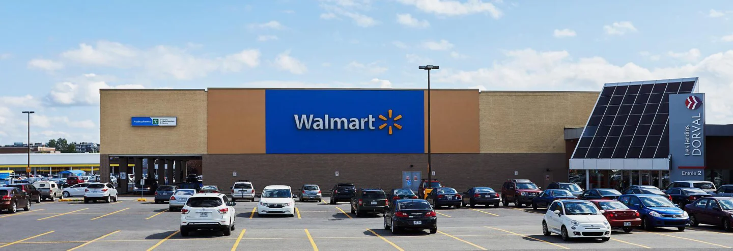 Wide-angle view of the Walmart store at Les Jardins Dorval shopping centre. The store facade features a large blue and orange color block with the Walmart logo prominently displayed in the center. To the right, a modern glass-panel entrance with a tall sign reads “Les Jardins Dorval - Entrée 2.” The parking lot in the foreground is filled with a variety of cars, and the sky above is partly cloudy.
